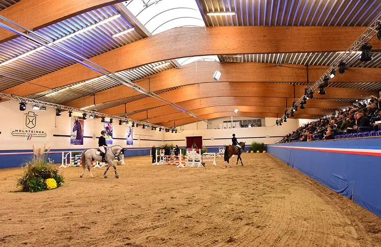 Fritz-Thiedemann-Halle im Veranstaltungsbetrieb: geschwungenes Holzdach, Lichttraversen, voll besetzte Tribüne und Reiter auf der Sandbahn